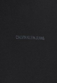 Tejido negro con una textura suave y un logo reflectante que dice "CALVIN KLEIN JEANS" sutilmente grabado. Diseño simple y minimalista.