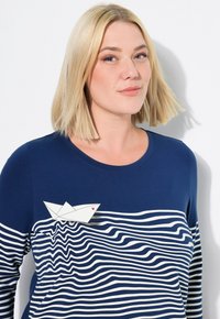 Tummansininen pitkähihainen paita, jossa on valkoisia vaakasuoria raitoja ja paperivene-design rintamuksessa, johon on lisätty pieni punainen sydän.
