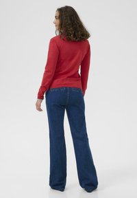 Piros, kötött pulóver, amely testhezálló fazonú és kerek nyakú, kék trapéz farmernadrággal párosítva. A pulóver alján kör alakú, texturált minta található.