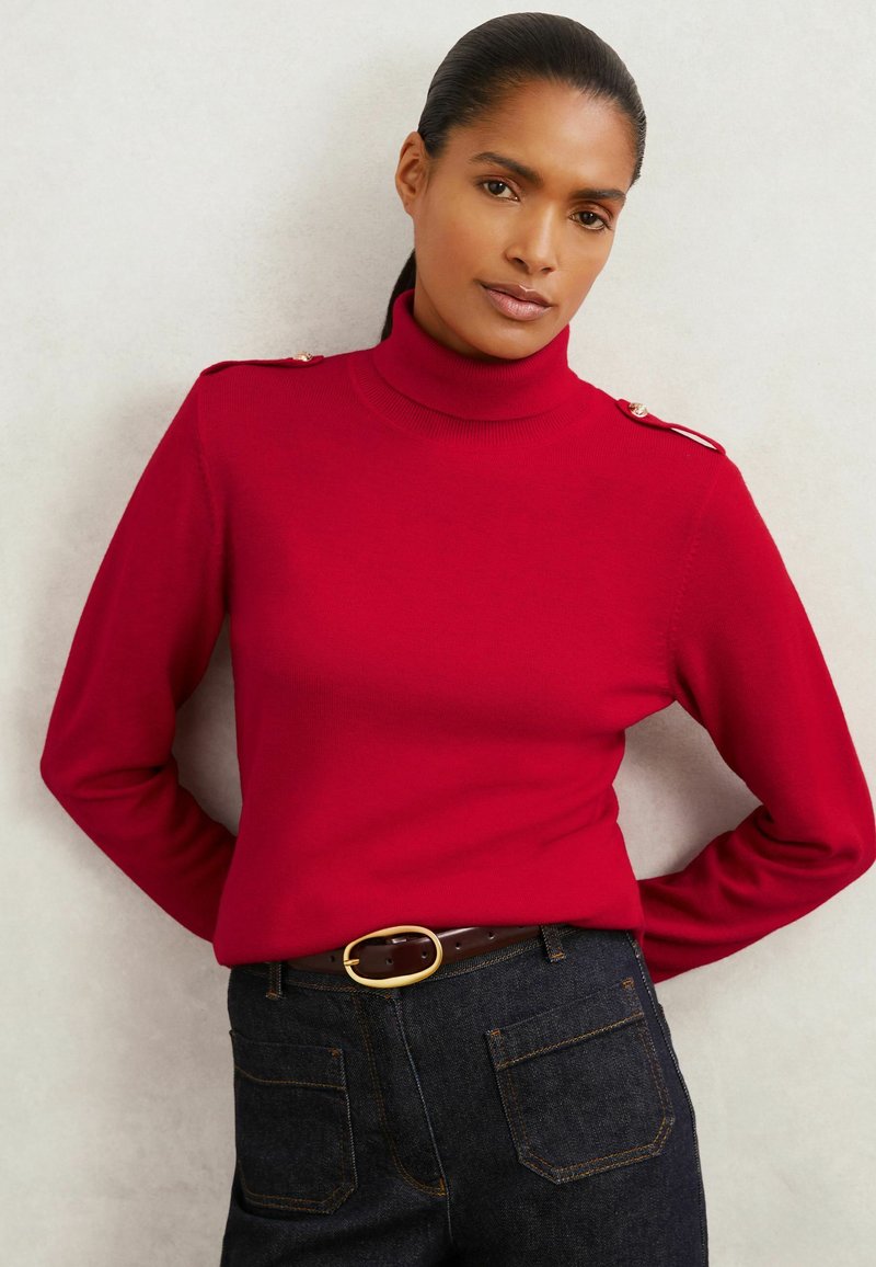 Maglione rosso a collo alto con maniche lunghe, caratterizzato da accenti in metallo dorato sulle spalle. Abbinato a jeans in denim scuro e una cintura marrone.