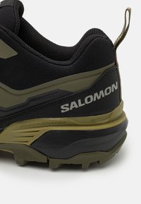 Črne in olivno zelene pohodne čevlje z mrežastim zgornjim delom, teksturiranim podplatom in "SALOMON" blagovno znamko. Vključuje napreden šasijski podrobnost na srednjem podplatu.