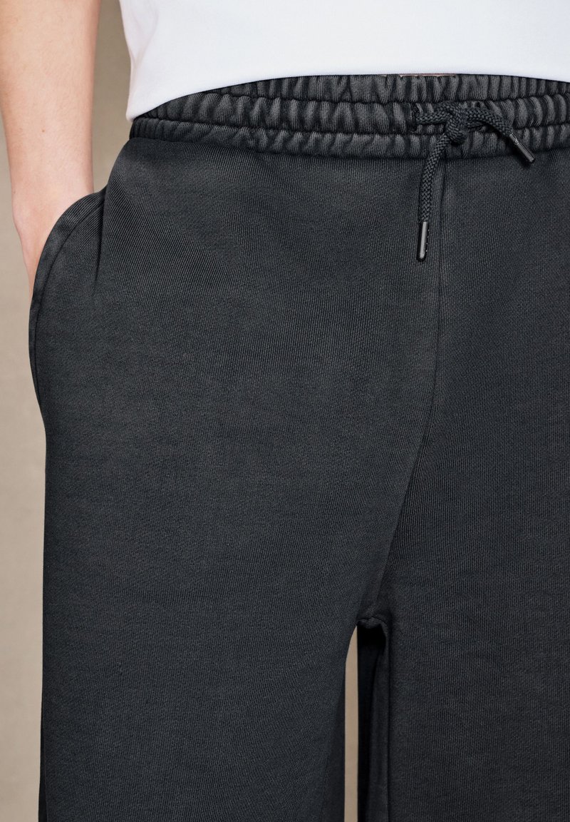 Gros plan d'une personne portant un pantalon de jogging noir à taille élastique avec cordon et la main dans la poche gauche.