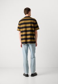 T-hirt met korte mouwen met horizontale zwarte en mosterdkleurige strepen, de stof heeft een gladde textuur. Draag het met vervaagde blauwe jeans en zwarte schoenen.