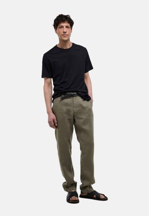 Homme debout portant un t-shirt noir à manches courtes, un pantalon vert olive avec une ceinture tissée et des sandales noires sur fond gris clair.