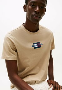 Camiseta de algodón beige con cuello redondo, que presenta un logo rectangular en azul marino, rojo y blanco con el texto "TOMMY HILFIGER" centrado en el pecho.