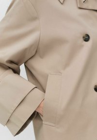 Beżowy płaszcz dwurzędowy z klapami, dużymi czarnymi guzikami i kieszenią naszywaną z boku. Gładki, teksturowany materiał.