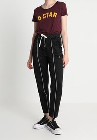 Camiseta marrón con "G-STAR" en amarillo, combinada con pantalones jogger negros con rayas laterales blancas y una cintura elástica. Zapatillas negras.