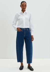 Weißes Langarmhemd mit Knöpfen, kombiniert mit lockeren blauen High-Waist-Jeans aus Denim. Das Model trägt schwarze Slipper.