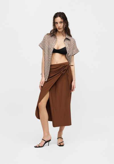 Γυναίκα με μαύρο crop top, πουκάμισο με σύντομο μανίκι και σχέδιο, καφέ wrap φούστα με πλευρικό σκίσιμο και μαύρα σανδάλια με τακούνι σε λευκό φόντο.