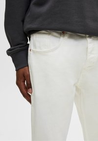 Jeans de denim blanco con un corte recto, detalle de costura y bolsillos frontales. Combinados con un sudadera gris oscuro en la cintura.