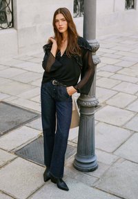 Blusa negra transparente con acentos de volantes, combinada con jeans de pierna ancha azul oscuro y botas de tobillo negras con punta, con un fondo de calle de piedra.