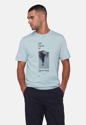 Homme aux cheveux courts portant un t-shirt bleu clair avec un graphisme noir et le texte "la main gauche de l'obscurité", mains dans les poches, regardant vers la gauche.