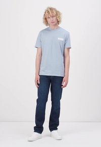 Hellblaues Baumwoll-T-Shirt mit kurzen Ärmeln, ausgestattet mit einem kleinen weißen Taschenlogo, kombiniert mit dunkelblauen Jeans und weißen Sneakers.