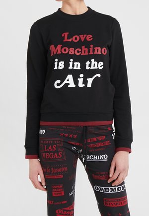Modèle portant un sweat-shirt noir Moschino avec l’inscription « Love Moschino is in the air » et un pantalon assorti à imprimé voyage.
