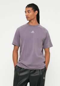 Vyras su ilgais pintais plaukais, vilkintis purpurinę Adidas trumpomis rankovėmis marškinėlius ir juodas Adidas kelnes, stovi su rankomis kišenėse, žiūrintis į kairę.