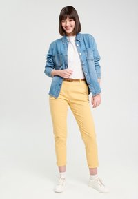 Chemise en jean bleu clair à boutons, t-shirt blanc et pantalon jaune court. Baskets blanches. Fond minimal et neutre.