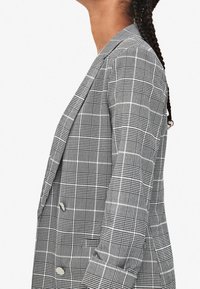 Šedý kostkovaný blazer s dvouřadým designem, klopovým límcem a dvěma velkými knoflíky, s texturovanou tkaninou a strukturovaným siluetou.