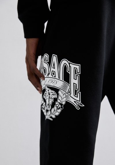 Čierne jogger nohavice z látky s bielym logom "Versace", ktoré obsahujú grafiku Medúzy a vavrínové akcenty. Hladká textúra a ležérny dizajn.