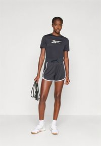 Juoda sportinė marškinėliai su balta logotipu, derinami su juodais šortais, turinčiais baltą apvadą. Modelis laiko šokdynę ir dėvi baltus sportinius batelius.