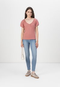 Top roz cu mâneci bufante, decolteu în V, din material texturat, combinat cu blugi skinny de culoare albastră deschisă și adidași bej, ținând o geantă mică albă.