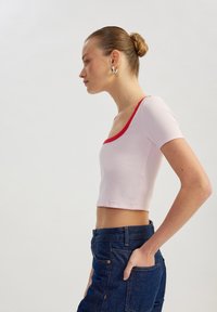 Lys pink ribbet crop top med korte ærmer og en rød halsudskæring. Modellen kombinerer det med mørke denim jeans, hænderne i lommerne, set fra siden.