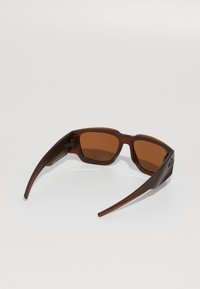 Brune solbriller med mat finish, rektangulære linser og slanke, buede stænger. Linserne er tonet brune. Bemærkelsesværdigt logo på siden.