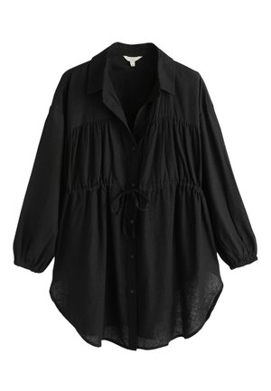 Femme aux cheveux en chignon bas portant une robe noire ample à manches longues, debout près d’un arbre et des plantes avec une lumière douce et des ombres.