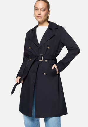 Femme portant un trench-coat noir ceinturé par-dessus un haut blanc et un jean bleu, ajustant la ceinture du manteau avec une main dans la poche.