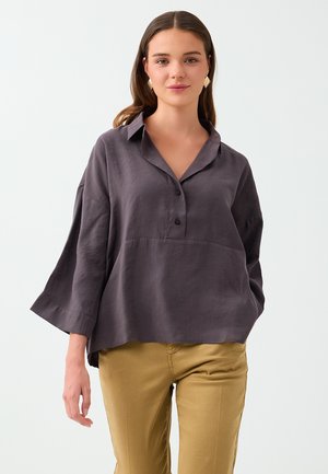 Femme portant un chemisier ample gris foncé boutonné avec des manches larges et un pantalon jaune moutarde sur un fond clair.