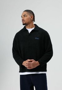 Schwarzes, strukturiertes Fleece-Poloshirt mit Kragen, verziert mit blauem, gesticktem Branding. Modell trägt es über einem weißen Hemd und dunklen Jeans.