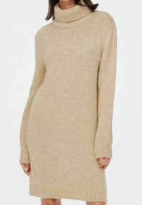 Femme portant une robe pull beige en tricot avec un col roulé et des manches longues sur un fond blanc uni.