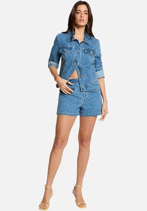 Kvinde iført en mønstret blå denimjakke med opkærede ærmer og matchende shorts, kombineret med beige højhælede sandaler på hvid baggrund.