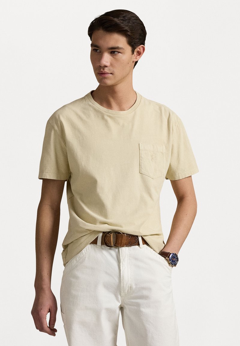 Beige, rövid ujjú póló, mellzsebbe varrott zsebbel, fehér nadrággal és barna övvel párosítva. A modellen sötét haj és óra van.