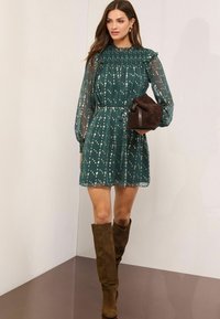 Zelené kvetinové mini šaty s priesvitnými dlhými rukávmi, volánikovým výstrihom a pásom stiahnutým v páse. Skombinované s hnedými semišovými čižmami po kolená a hnedou taškou.