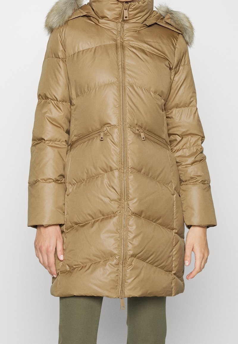 Kobieta ubrana w beżową pikowaną puchową kurtkę z kapturem obszytym futrem, zapinaną na zamek z przodu i posiadającą zapinane na zamek boczne kieszenie, stojąca na jednolitym tle.