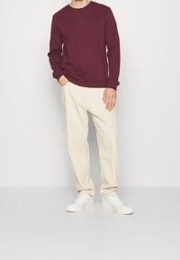 Bordowa bluza z okrągłym dekoltem, zestawiona z jasnobeżowymi, zwężanymi spodniami i białymi trampkami, prezentująca luźny krój i gładką fakturę.