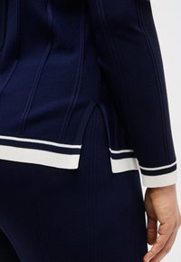 Κοντινό πλάνο πουλόβερ σε σκούρο μπλε με ριμπ ύφανση και φούστα με λευκή ρίγα στην άκρη και σχίσιμο στο πλάι, δείχνοντας μέρος του χεριού και του καρπού του ατόμου που το φοράει.