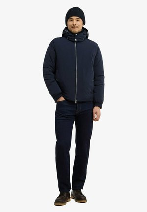 Homme portant une doudoune bleu marine, un bonnet noir, un jean foncé et des chaussures marron, debout avec une main dans la poche sur fond blanc.