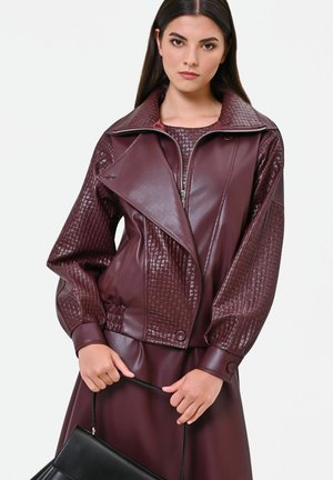 Donna che indossa una giacca di pelle bordeaux con dettagli trapuntati e un vestito abbinato, che tiene in mano una borsa nera su sfondo bianco.