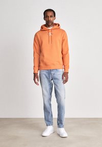 Felpa arancione con cappucci e logo, abbinata a jeans blu chiaro e sneaker bianche. Design semplice, materiale in cotone, vestibilità rilassata.