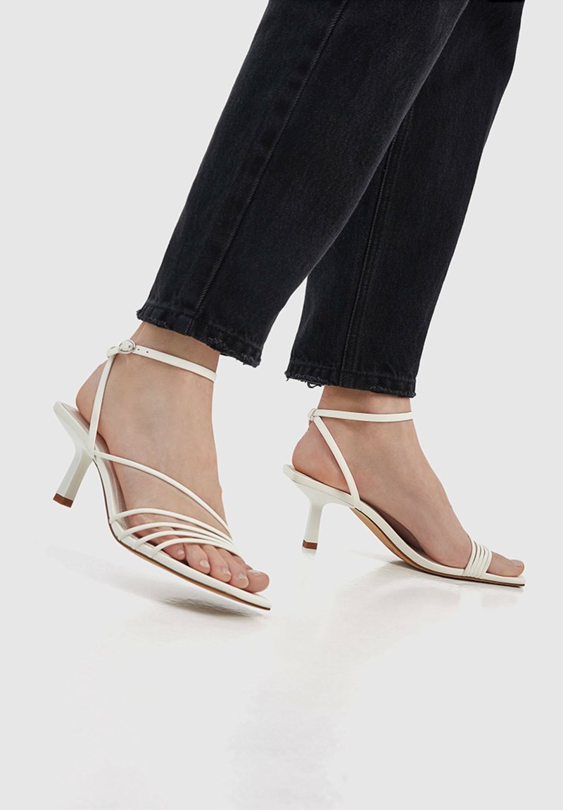 Pieds portant des sandales blanches à talons hauts et fines brides, associées à un jean noir coupé au-dessus de la cheville, sur fond uni.