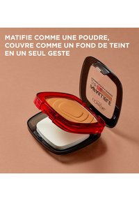 Poudre compacte avec un boîtier rouge et noir, contenant une poudre beige et une éponge applicatrice blanche à l'intérieur. Logo du produit gravé visible.