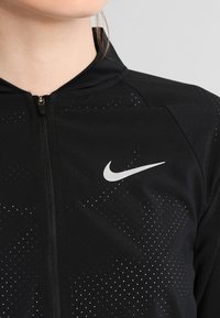 Černá bunda Nike se zipovým uzávěrem, která má jemný tečkovaný vzor a bílé logo na přední straně. Materiál se zdá být hladký a lehký.