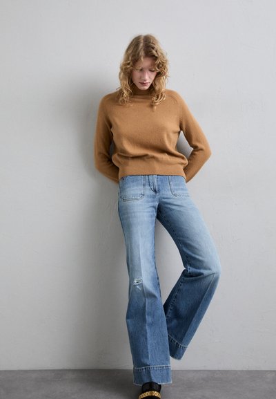 Világosbarna kötött pulóver magas nyakkal, világoskék, bő szárú farmernadrággal, melynek térden van egy kicsit sérült része, semleges háttér előtt.