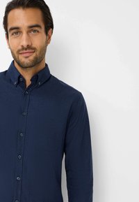 Camicia button-up blu navy con tessuto strutturato, maniche lunghe e taschino sul petto. Presenta un colletto classico e bottoni abbinati.
