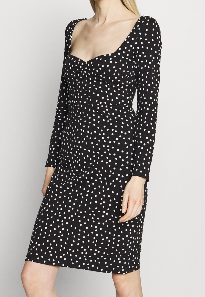 Femme portant une robe noire à manches longues avec des pois blancs et un décolleté en cœur, debout devant un fond uni.