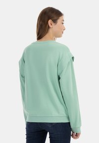 Felpa verde menta con una vestibilità rilassata, scollatura rotonda, maniche lunghe e polsini arrotolati, realizzata in tessuto morbido. Indossata con jeans blu scuro.