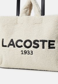 Borsa a spalla morbida di colore crema con ricami neri "LACOSTE" e "1933". Presenta manici neri corti e robusti e un tessuto texture.
