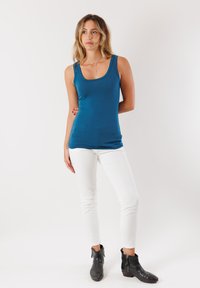 Canotta senza maniche di colore teal in uno stile aderente, abbinata a jeans skinny bianchi e stivali neri alla caviglia. Il tessuto appare liscio con una texture minimale.