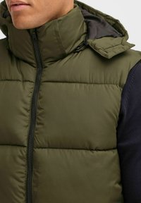 Olivgrön puffer-väst med hög krage, dragkedjestängning och horisontell sömnad. Har en svart dragkedja och huva med en slät, matt yta.